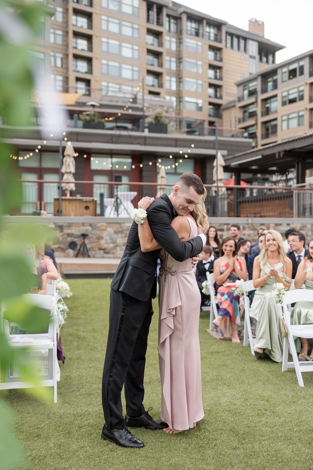 Wedding party celebrates outdoors on a grassy lawn with apartment buildings in the background and string lights overhead.
