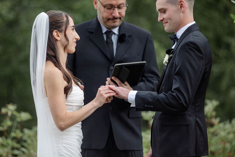 A couple exchanges rings during an outdoor wedding ceremony surrounded by greenery and natural light.