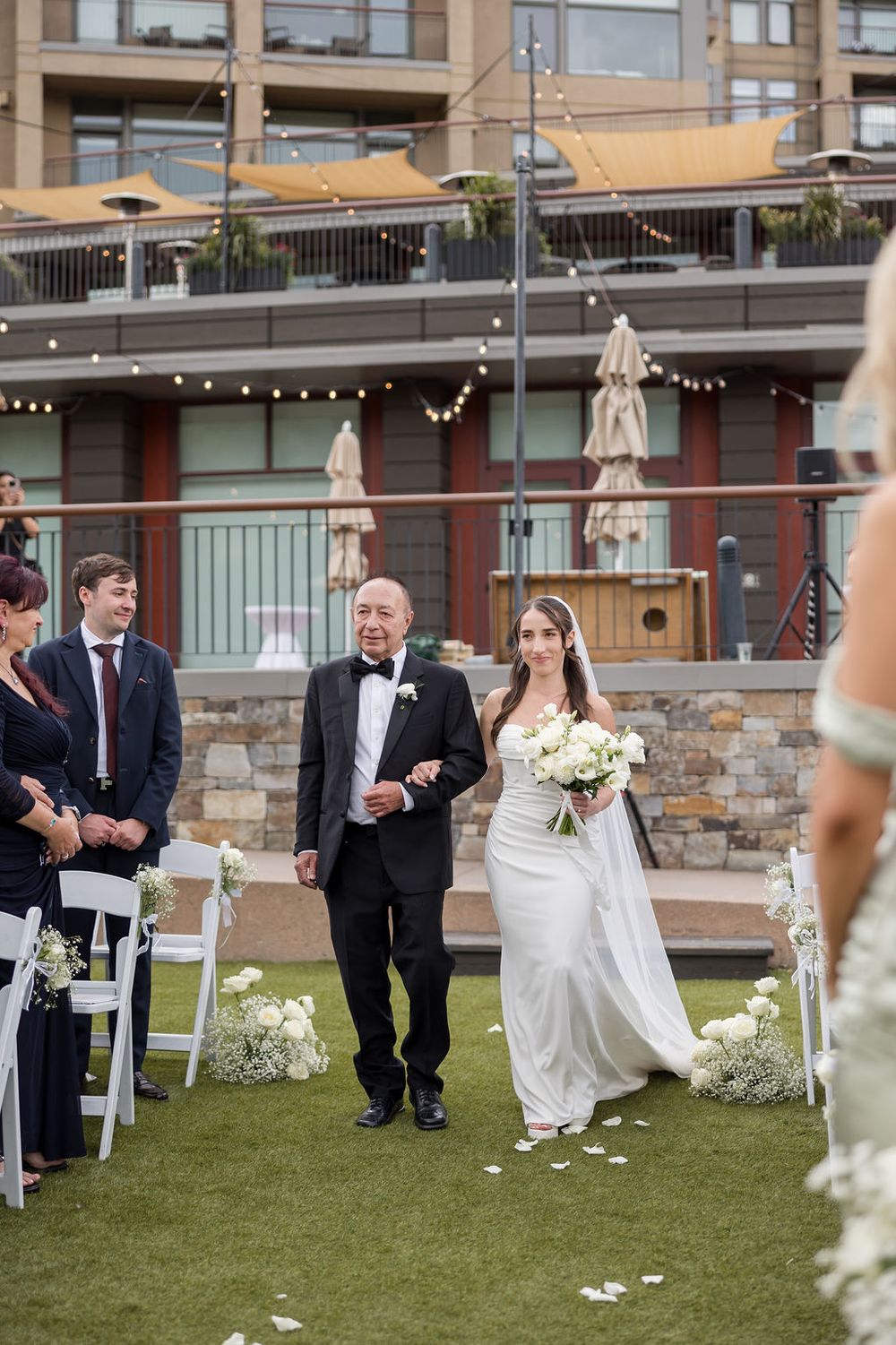 Wedding ceremony guests seated on white chairs on a grassy lawn with stone building and umbrellas in background.