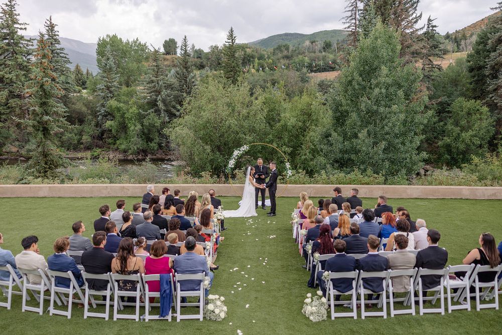 An outdoor wedding ceremony in a mountain setting with white chairs arranged on a grassy lawn surrounded by evergreen trees.