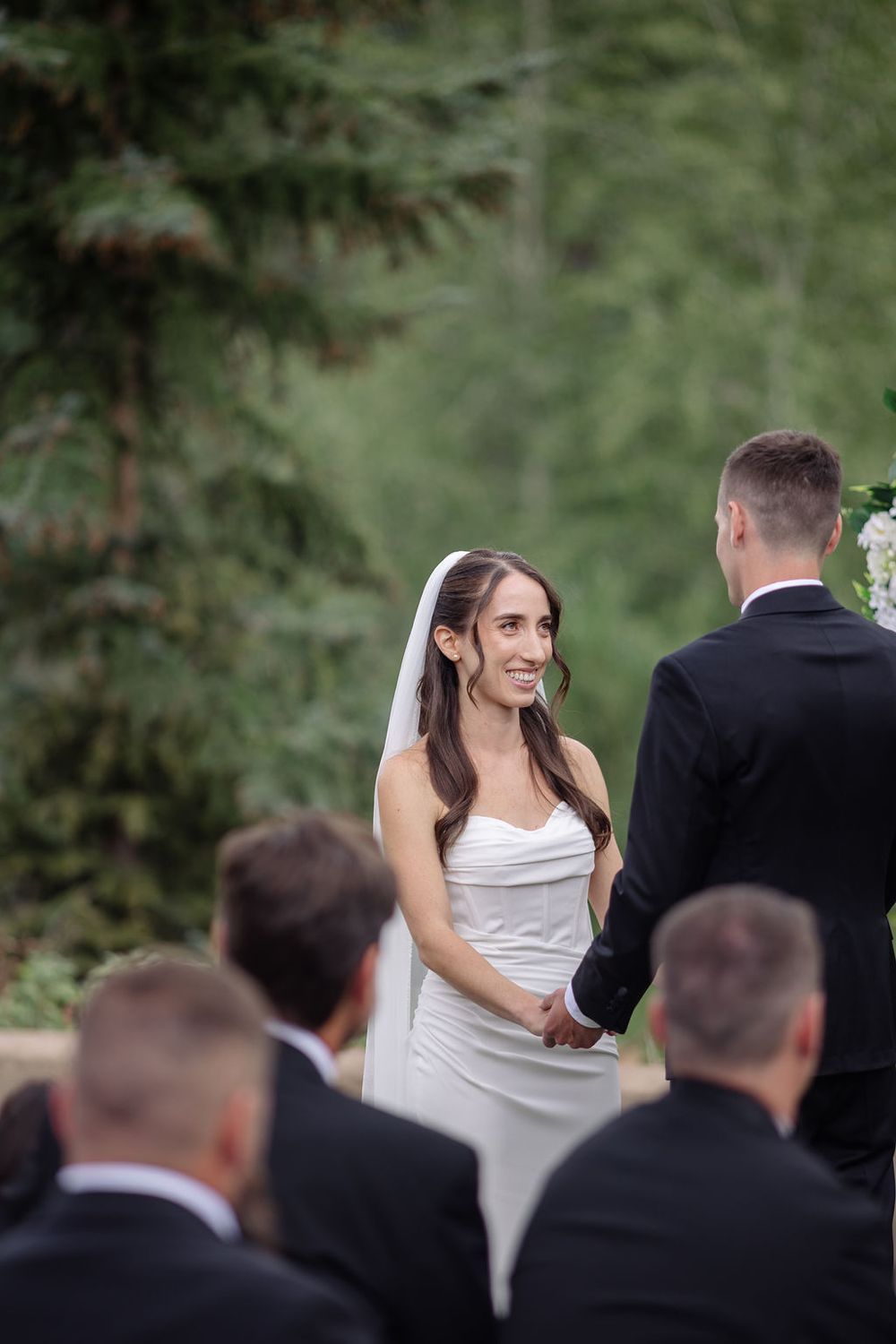 A wedding ceremony scene showing a bride in white dress and groom in black suit surrounded by guests in an outdoor setting.