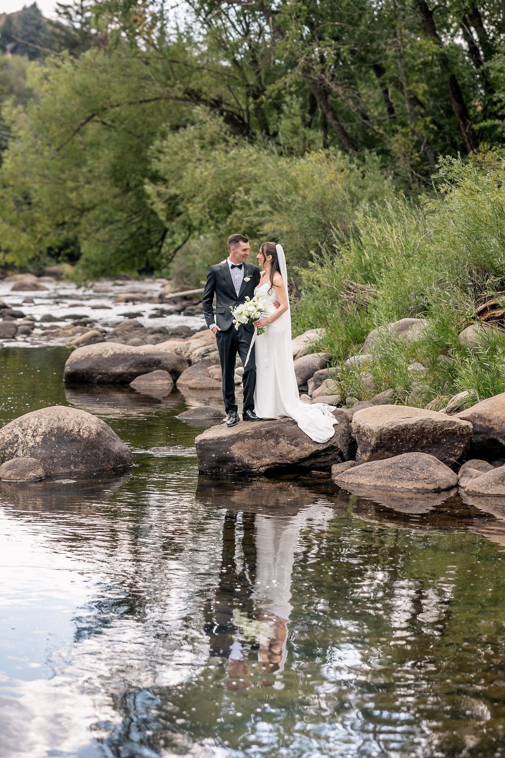 A couple in formal wedding attire poses together on large rocks beside a tranquil river in a scenic outdoor setting.