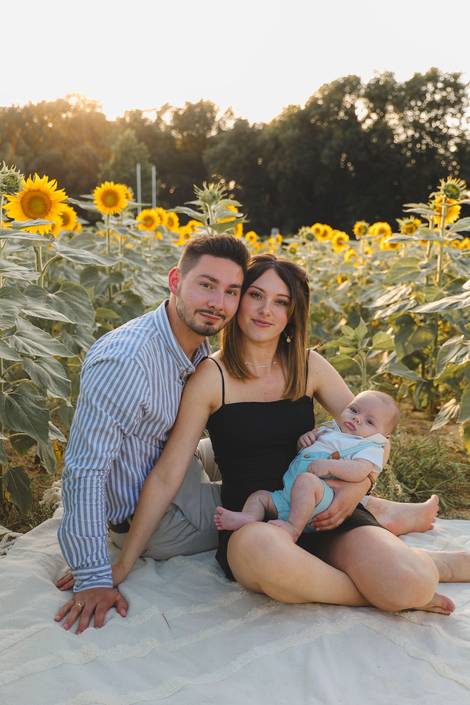 Young family with baby sitting together in sunflower field during golden hour sunset.