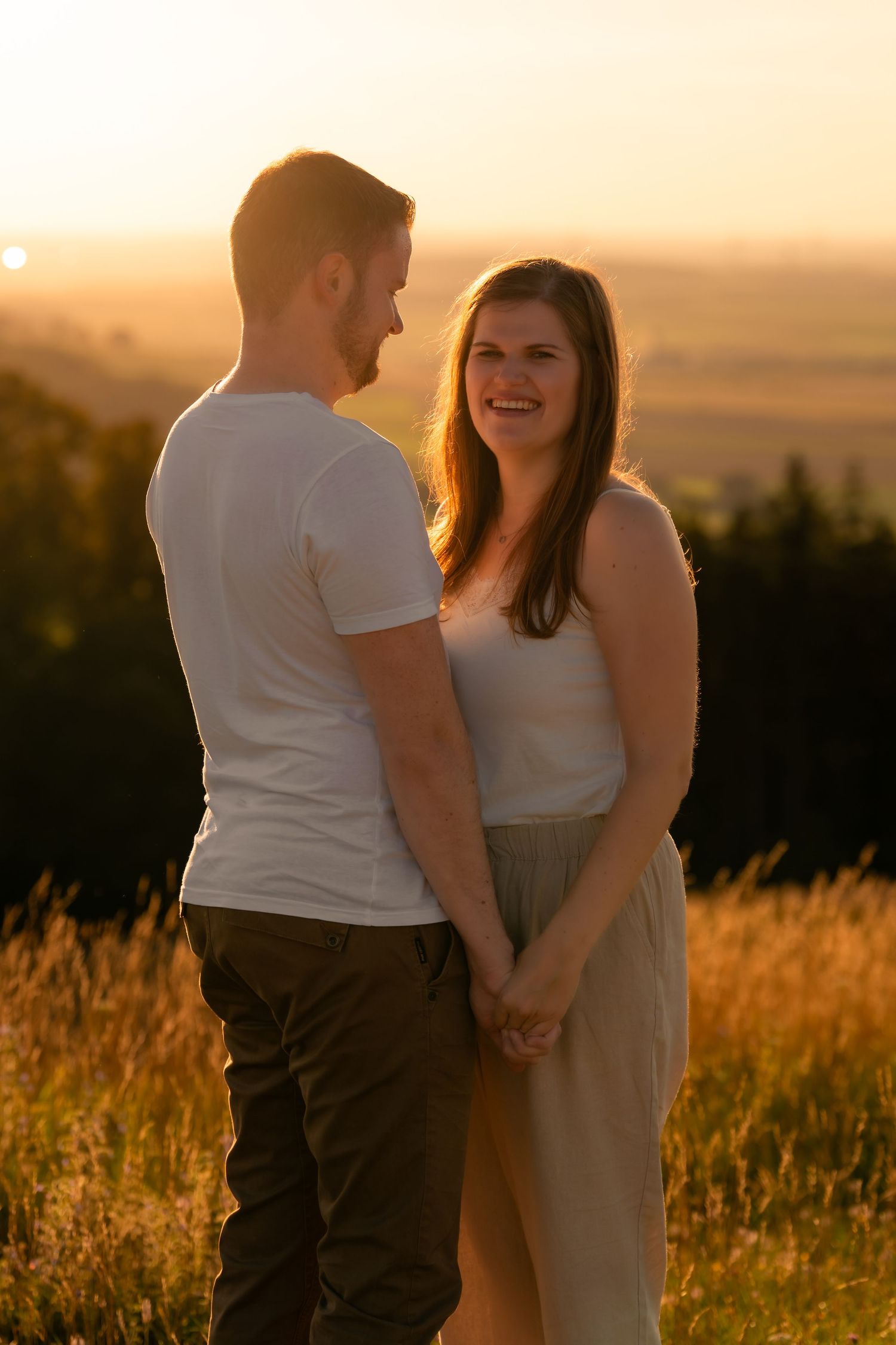 A couple embraces in a golden wheat field during sunset as warm light illuminates the romantic scene.