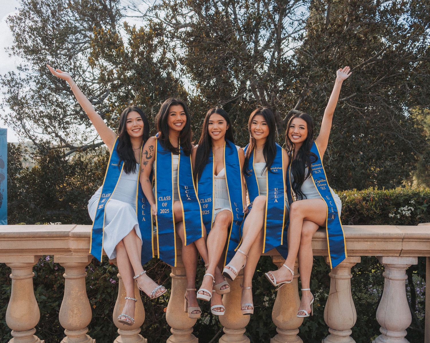 Five smiling female UCLA graduates in blue and gold stoles sit on a stone balustrade outdoors.