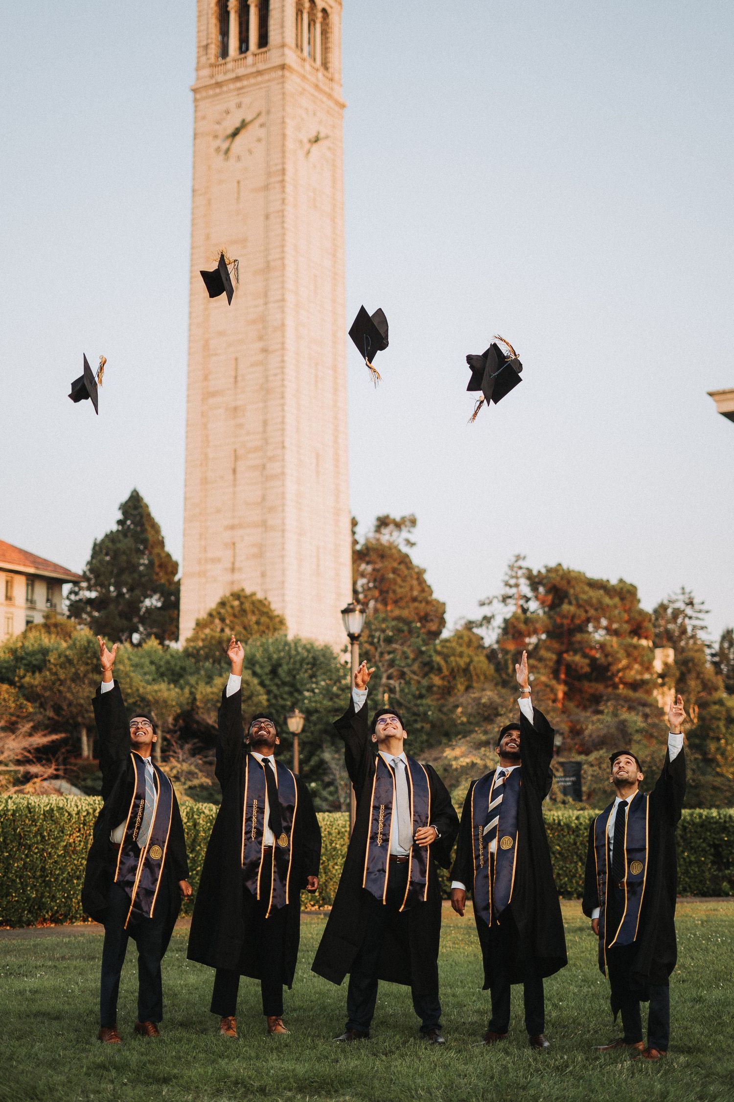 Five graduates in black robes tossing caps in the air near a tall clock tower on campus.