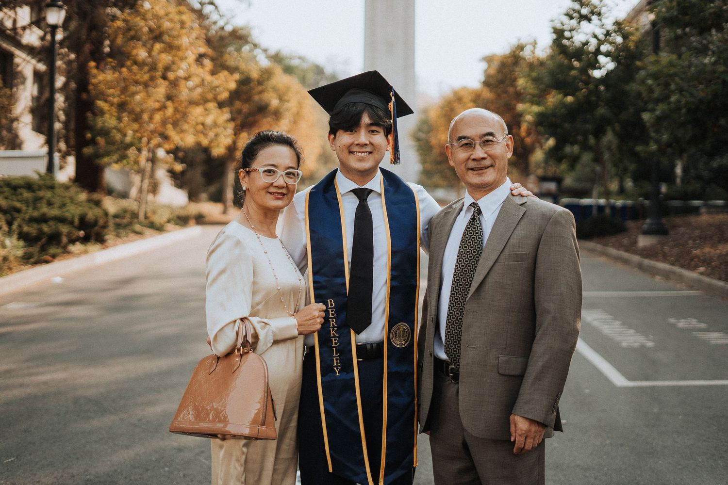 Proud graduate in cap and gown smiling with parents outdoors on a sunny university campus.
