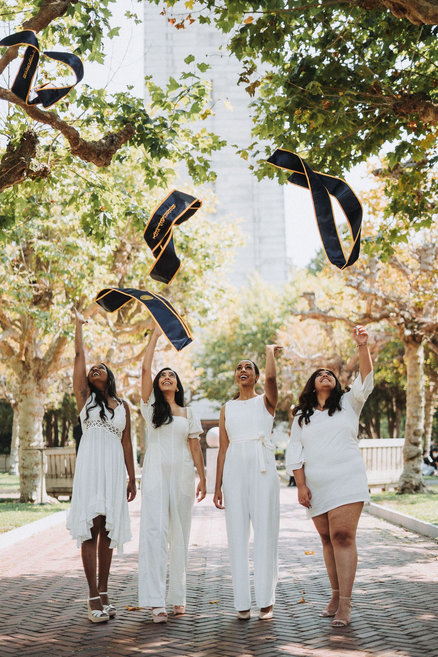 Four women in white outfits joyfully tossing graduation caps under tree-lined campus walkway.