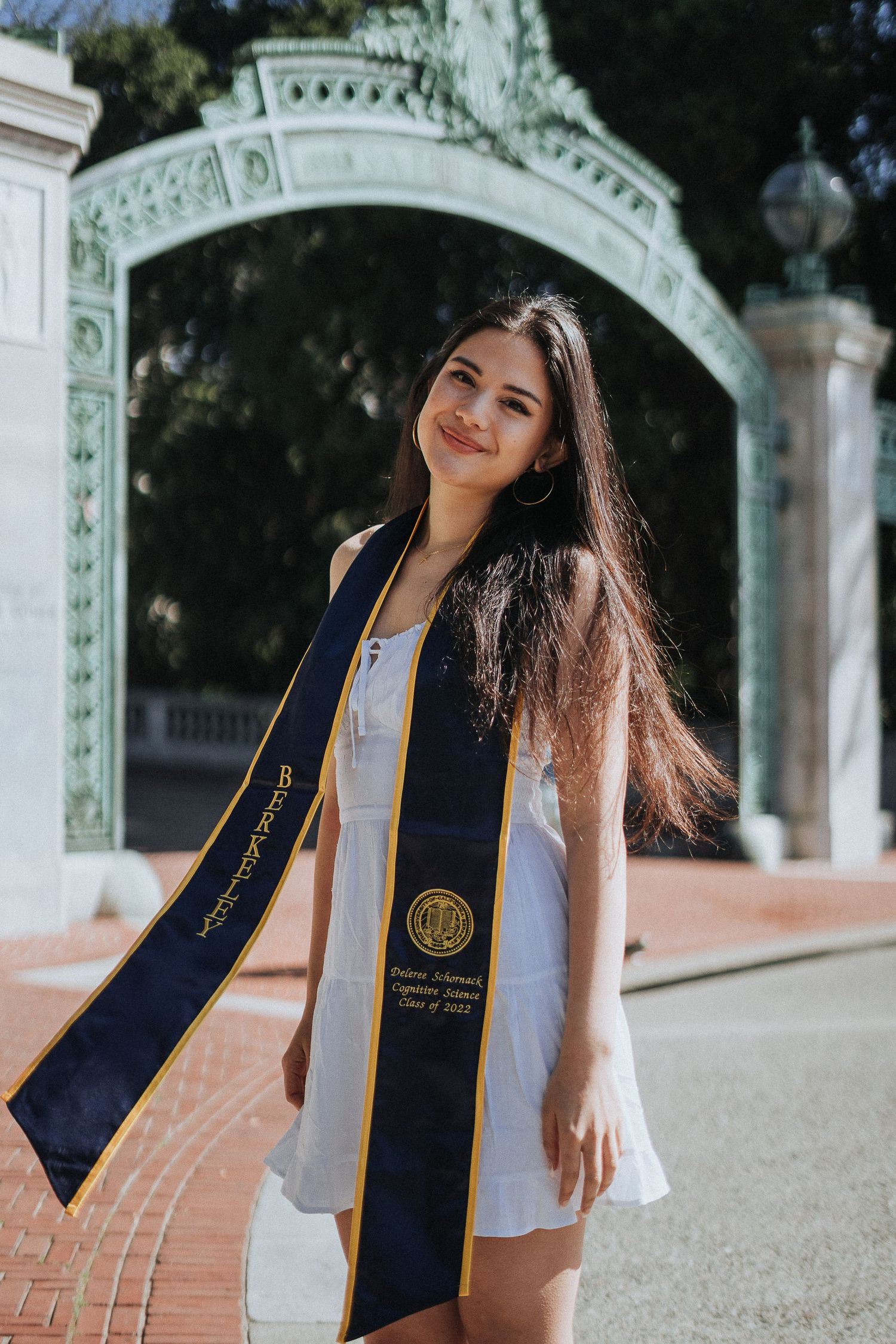 Smiling young woman in white dress wearing UC Berkeley graduation stole stands before Sather Gate.