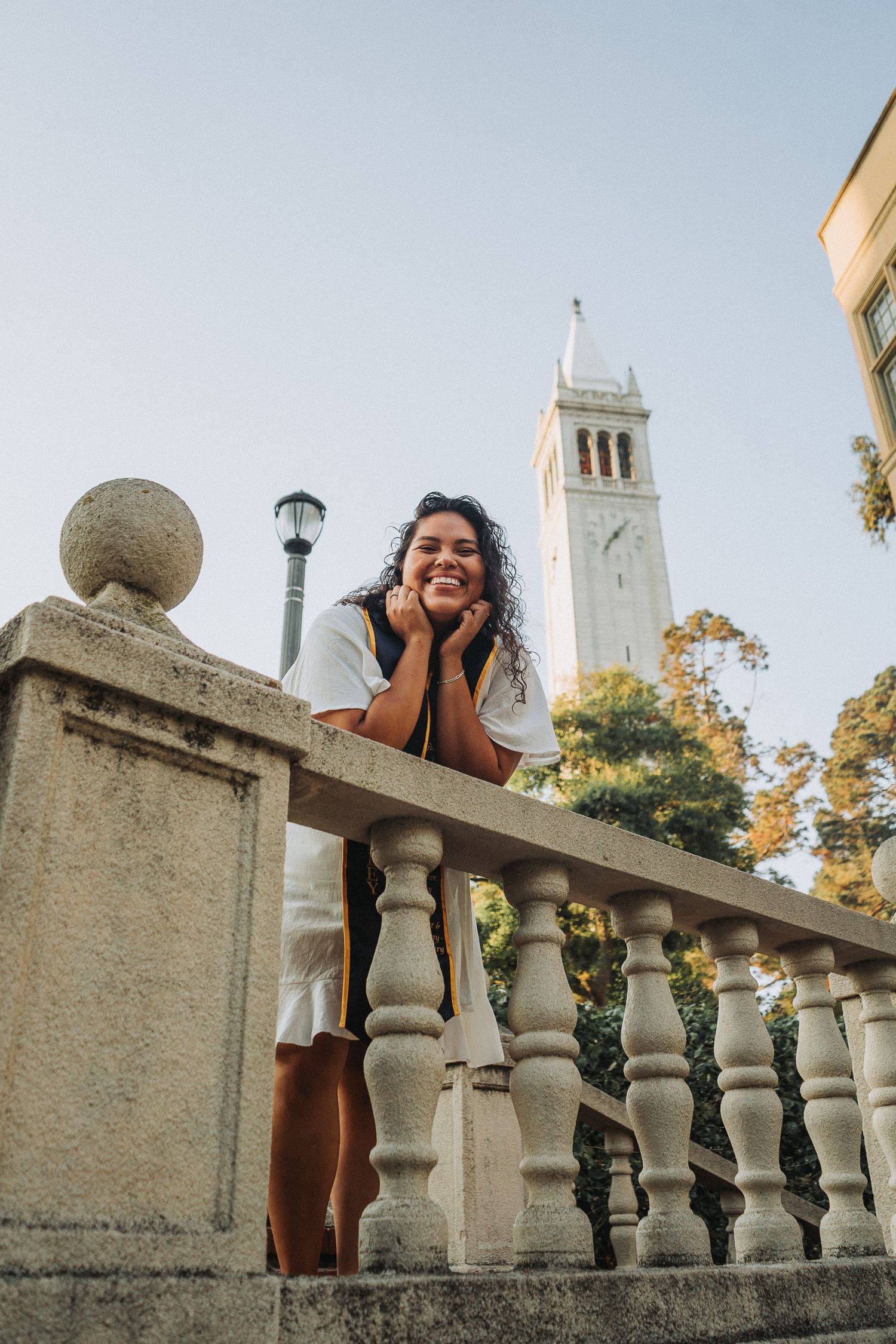 Smiling young woman leaning on stone balustrade with a university clock tower in the background.