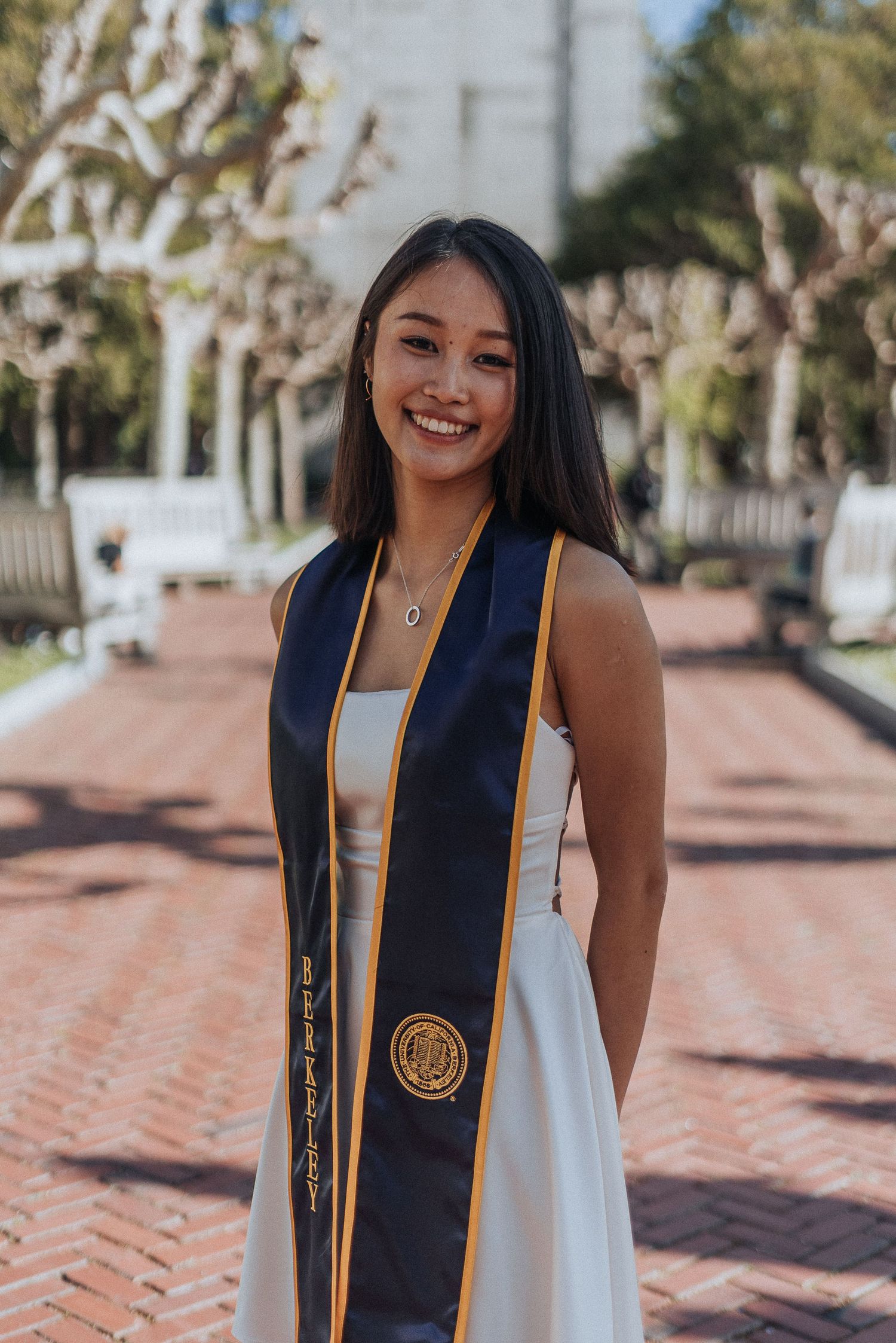 Smiling young woman in white dress and UC Berkeley navy graduation stole poses on a brick pathway.