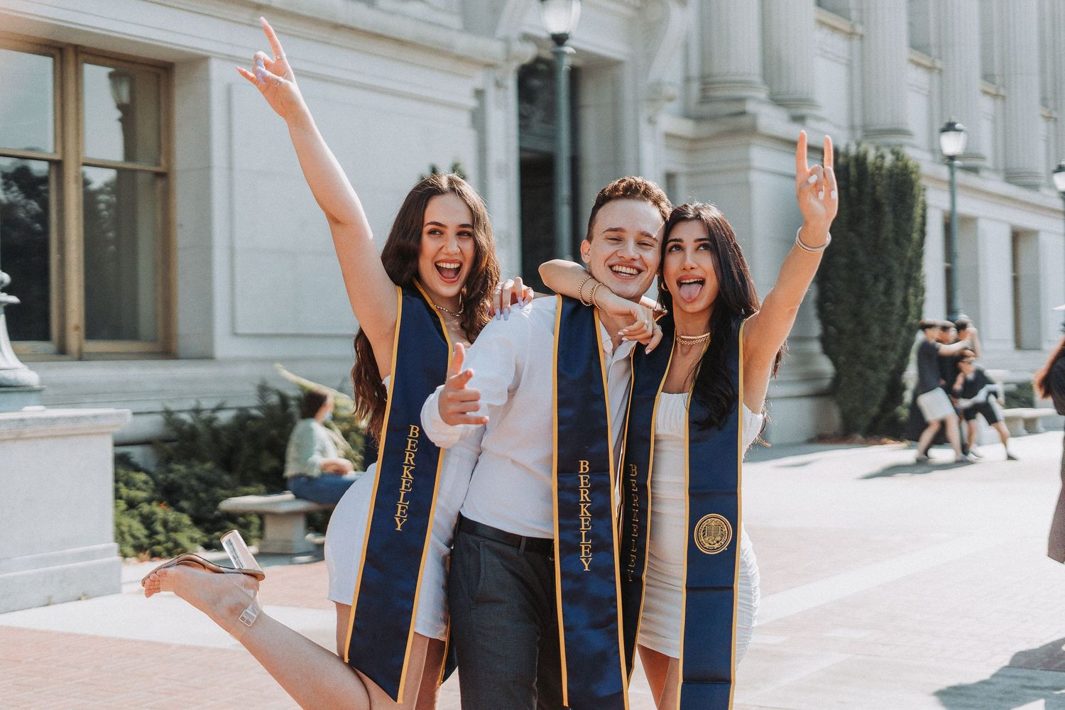 Three excited graduates in blue stoles celebrating outdoors in front of a university building.