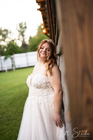 A bride in a white lace dress poses against a rustic wooden backdrop during a romantic outdoor wedding photoshoot.