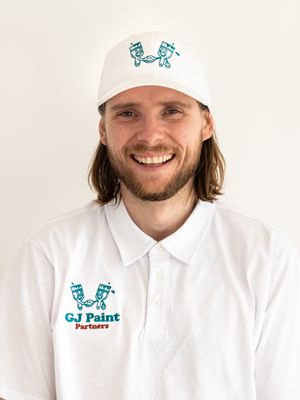 Professional painter wearing white uniform and branded cap with GJ Paint Partners logo.