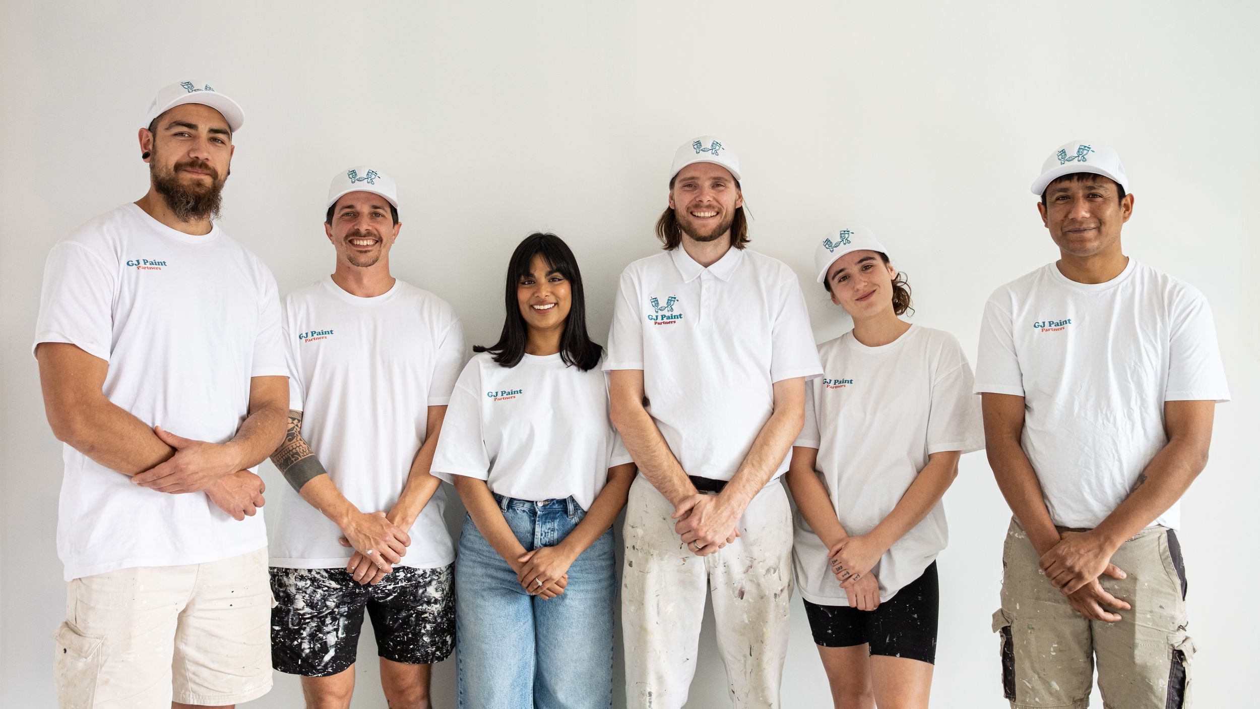A group of people wearing white t-shirts and jeans stand in a line against a white wall.