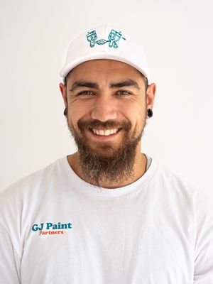 Painter wearing a white cap and t-shirt with company logo smiles warmly against light background.