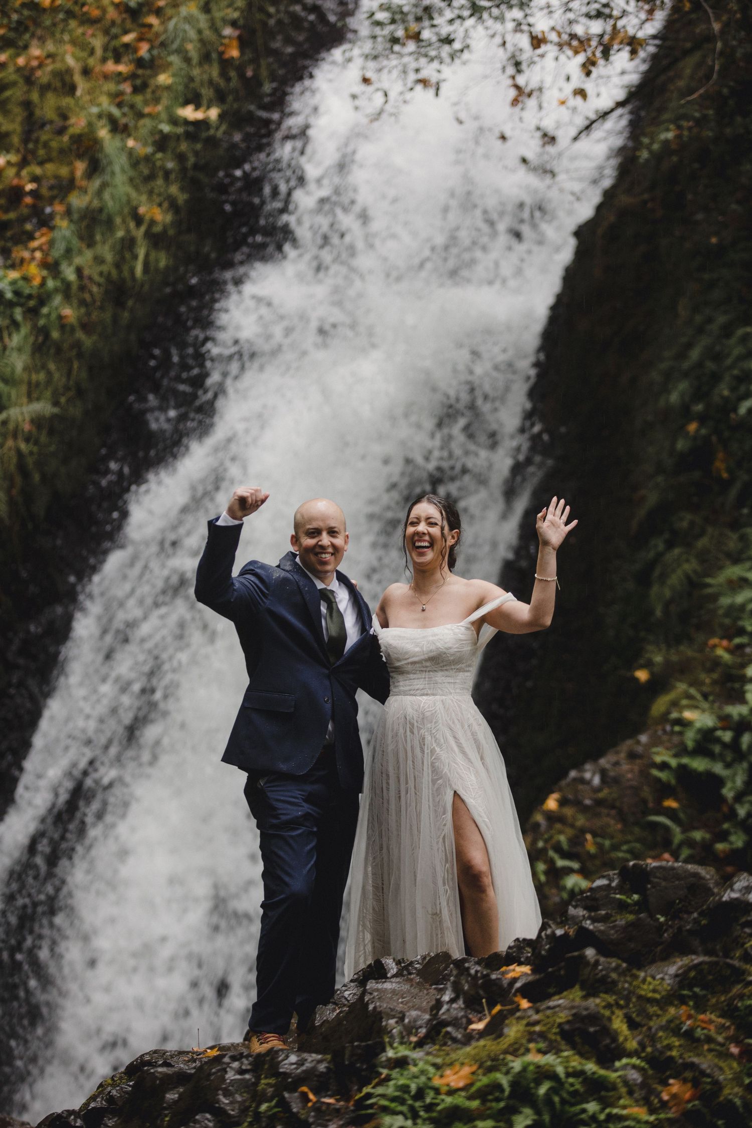 A couple in wedding attire poses together in front of a cascading waterfall surrounded by autumn foliage.