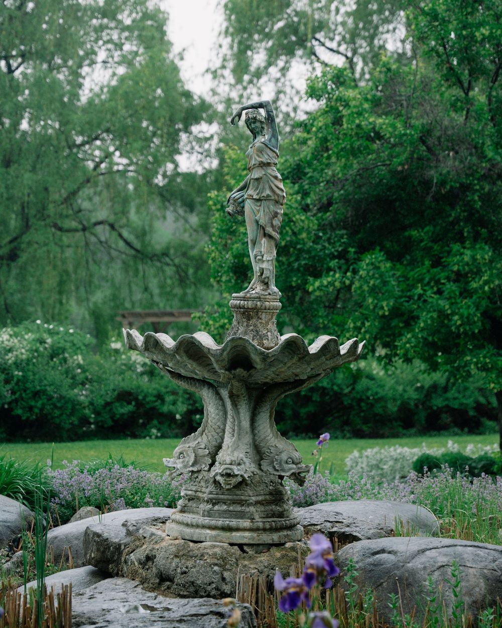 Stone garden fountain with tiered scalloped basins and classical female statue surrounded by lush greenery and purple iris flowers.