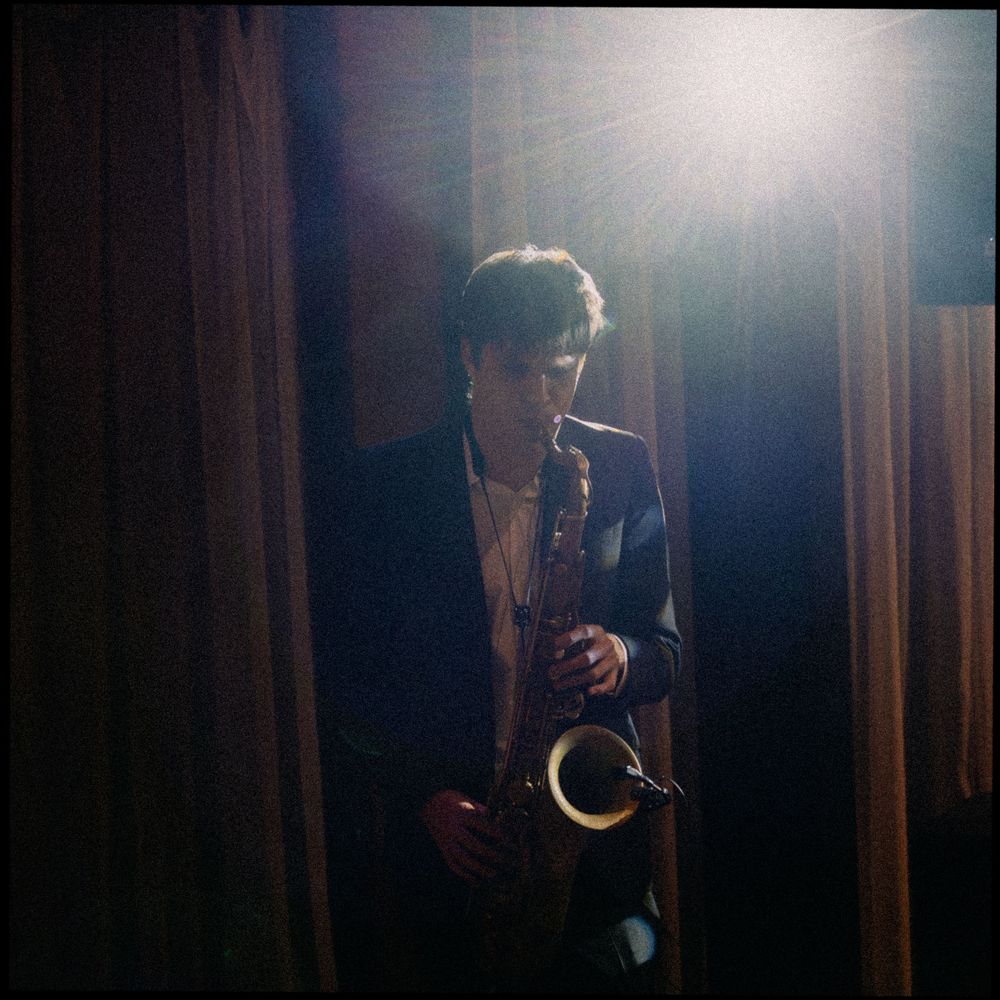 Musician plays saxophone dramatically backlit by bright light beside brown curtains at wedding.