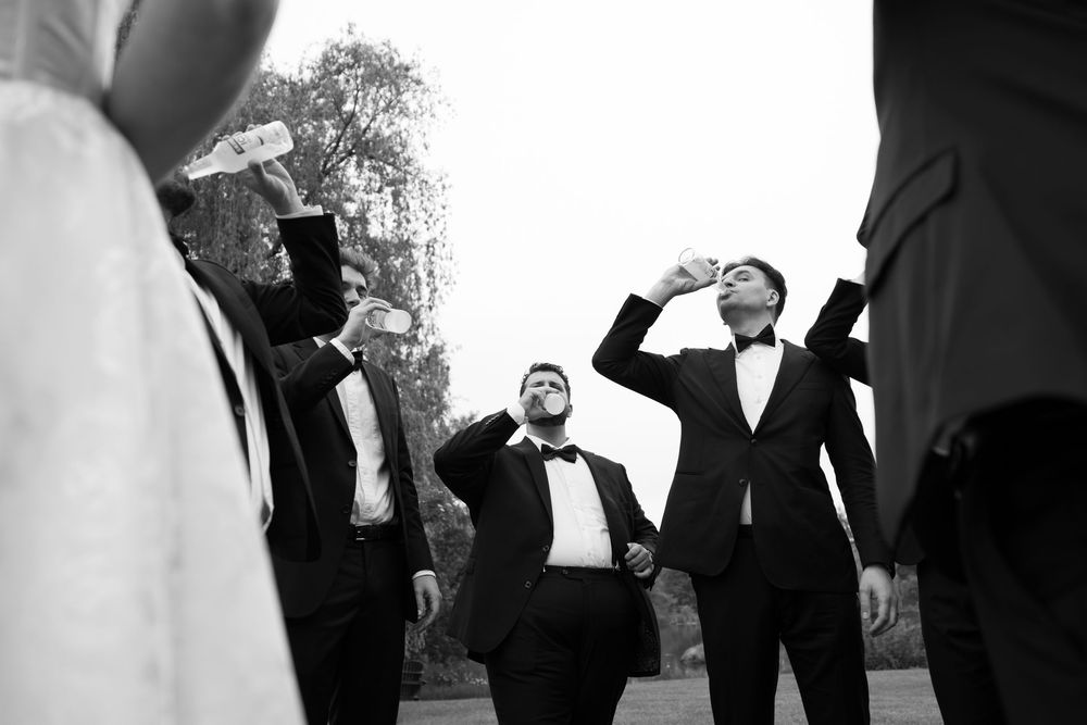 Black and white photo of groomsmen in tuxedos doing shots at a wedding celebration outdoors.
