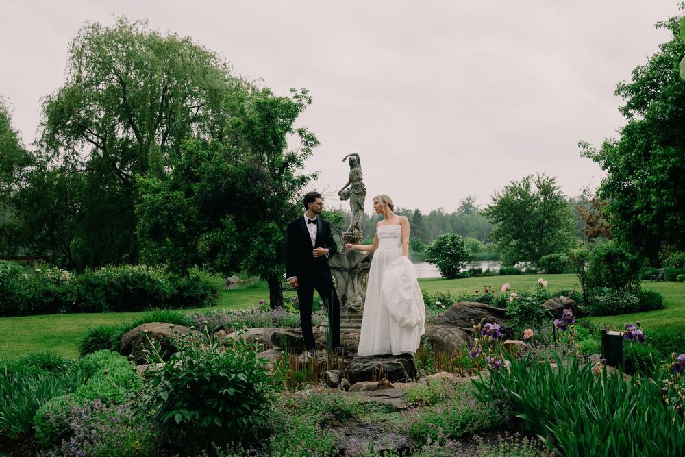 Bride and groom stand in a lush garden with a statue, surrounded by greenery on their wedding day.
