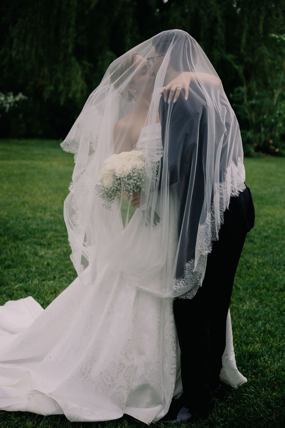Bride and groom sharing a romantic kiss under a lace wedding veil outdoors on green grass.