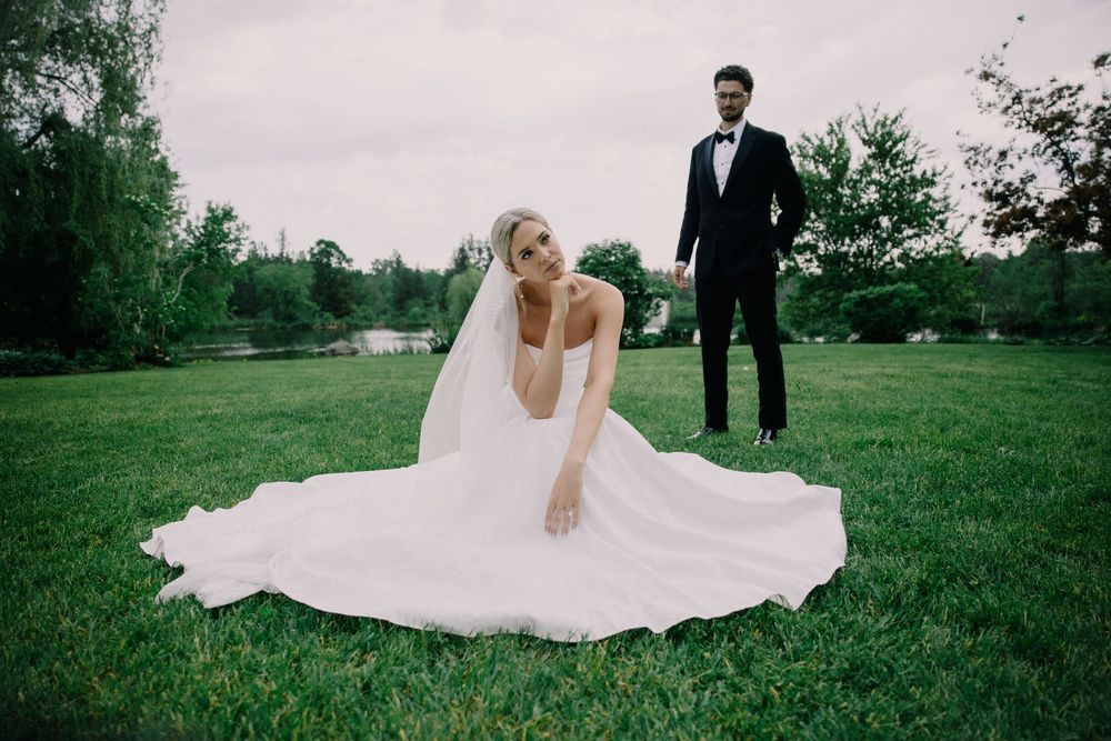 Bride in white gown sits on green grass while groom in black tuxedo stands behind her at outdoor wedding.