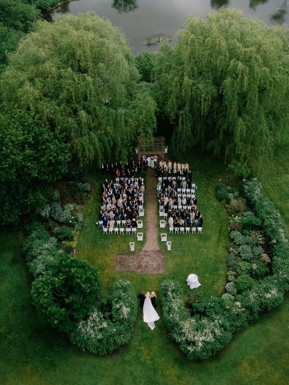 Aerial drone view of an elegant outdoor wedding ceremony surrounded by lush weeping willow trees and manicured gardens.