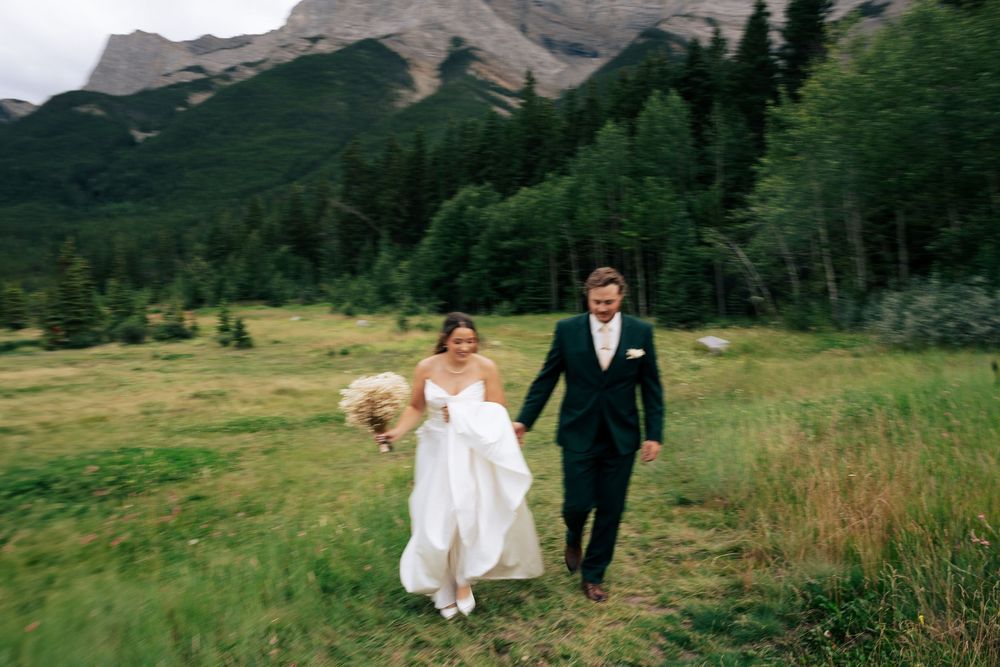 A couple walks through a scenic mountain meadow with evergreen trees and rugged peaks in the background.