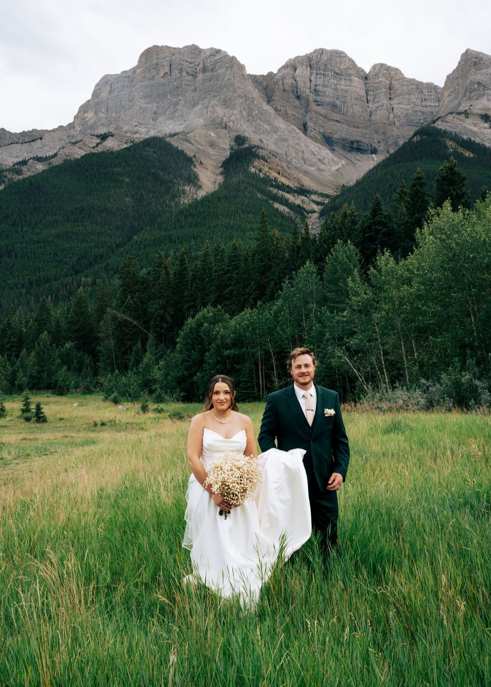 A couple in wedding attire walks through a mountain meadow with dramatic rocky peaks in the background.