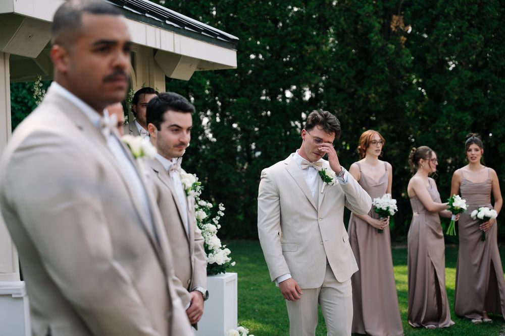 Wedding party in light beige and taupe attire poses outdoors on a sunny day with green trees in the background.