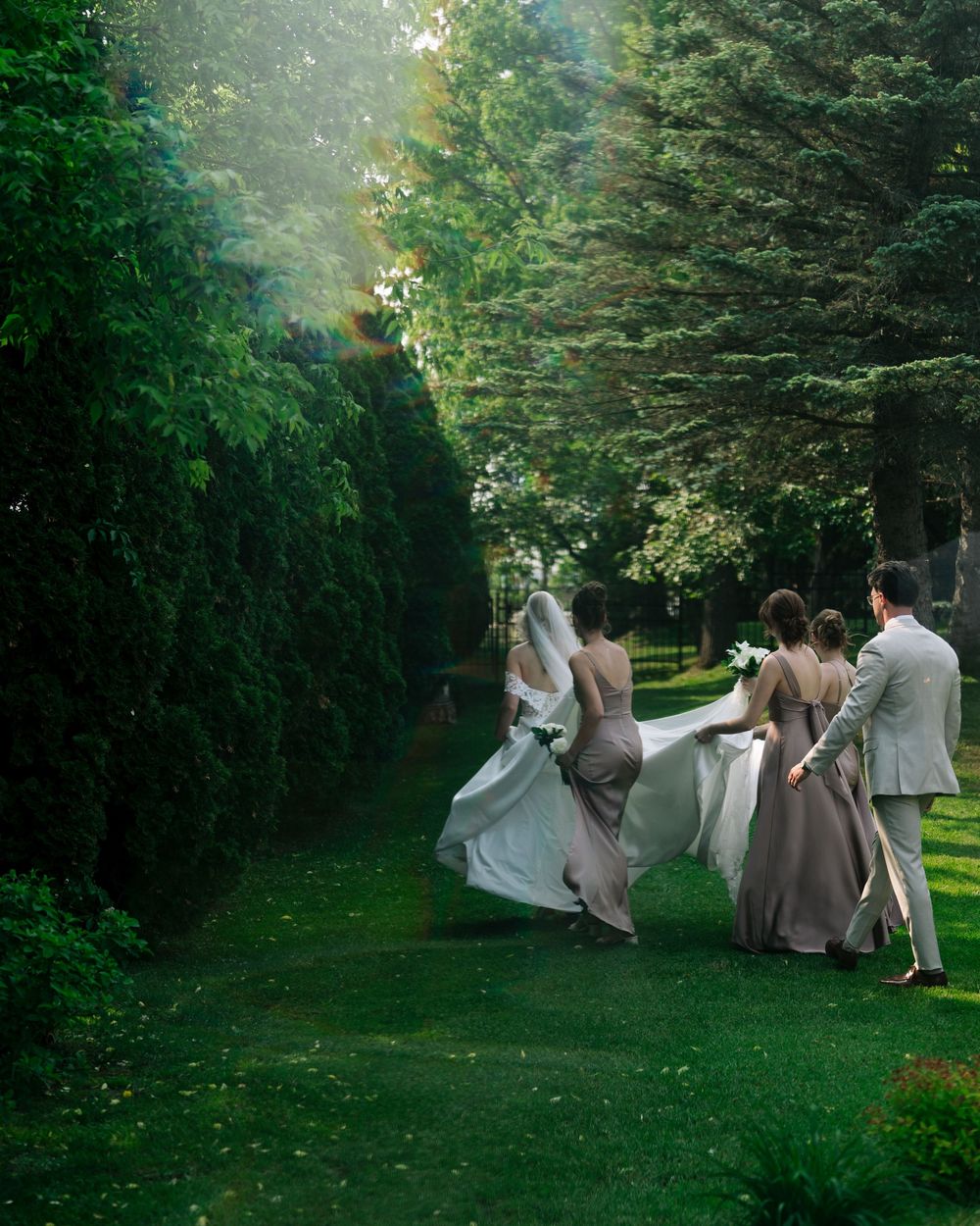Wedding party members playfully falling onto grass in a choreographed domino effect at an outdoor garden venue.