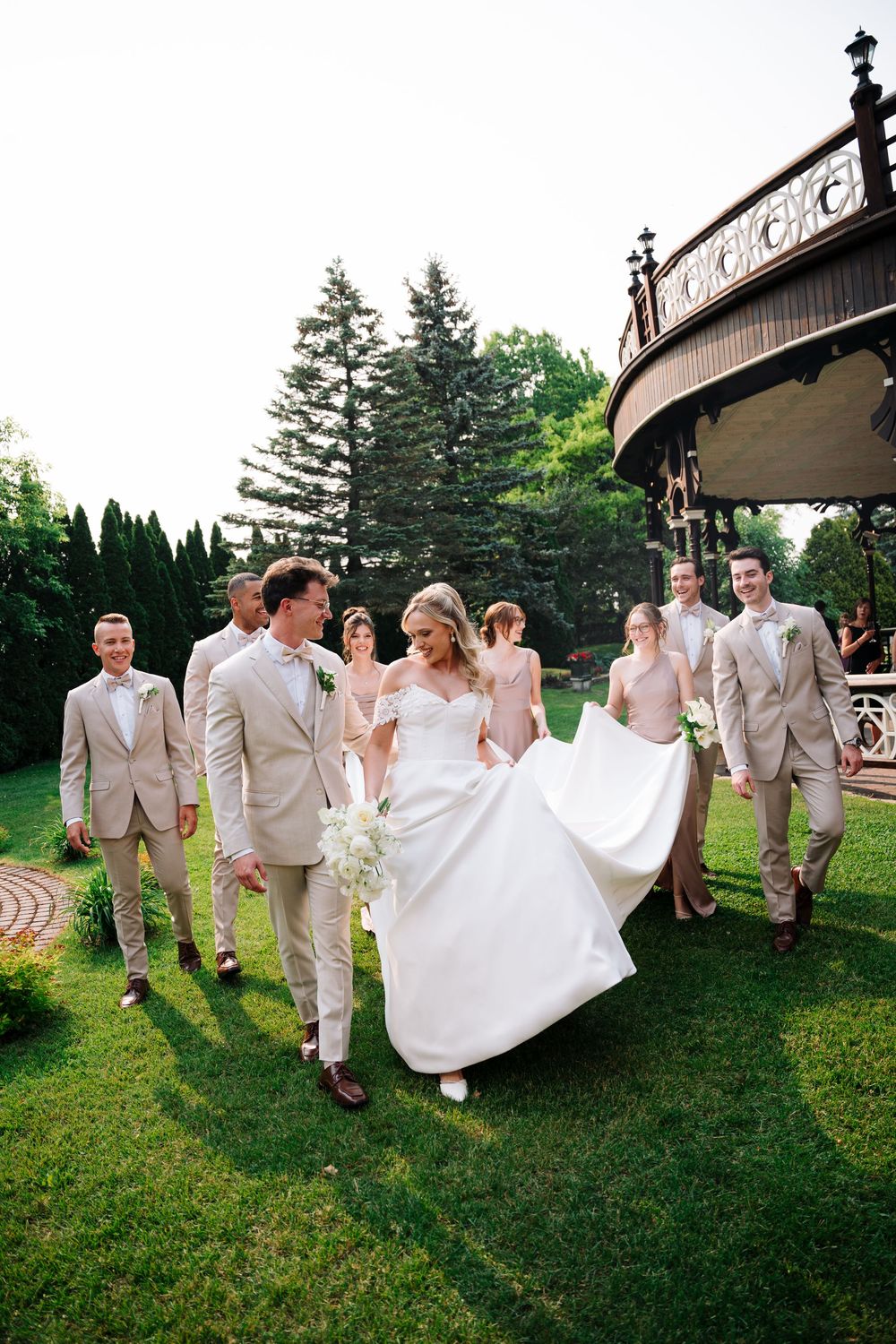 Wedding party in light colored attire walking together on grass with trees and buildings in the background.