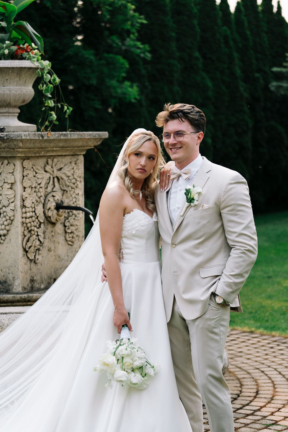 A couple poses in formal wedding attire near an ornate stone pillar in a garden setting.