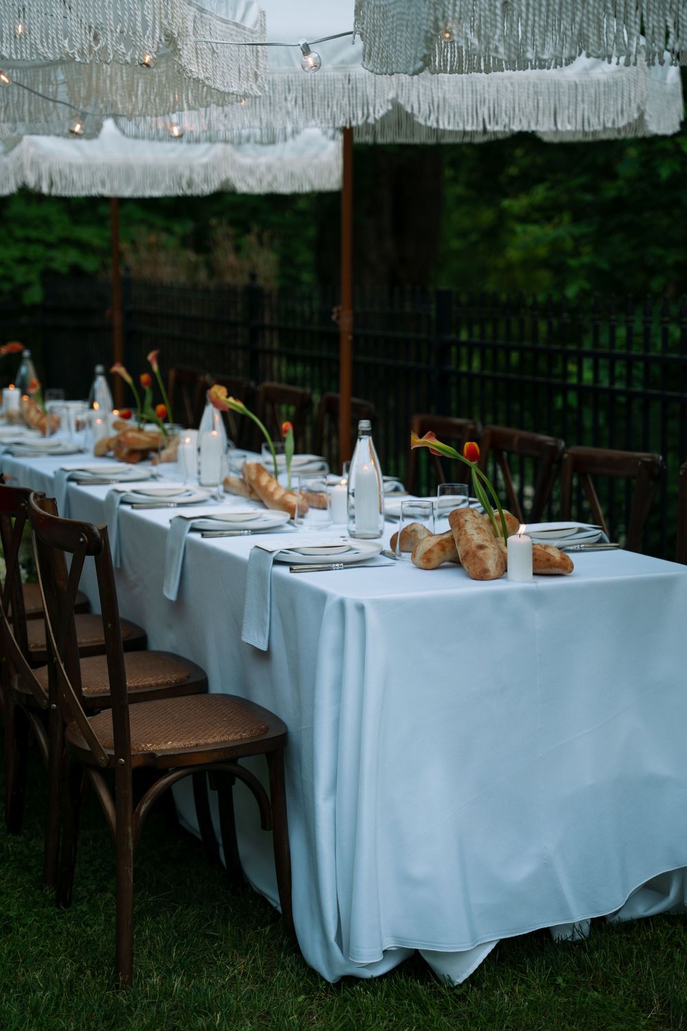 Long white tablecloth-covered tables set for an elegant outdoor dinner party with rustic wooden chairs and garden backdrop.