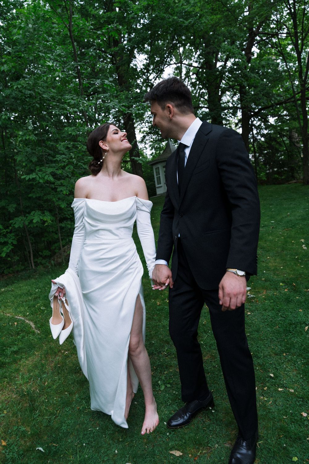 A couple in formal attire poses playfully on a green lawn surrounded by trees.