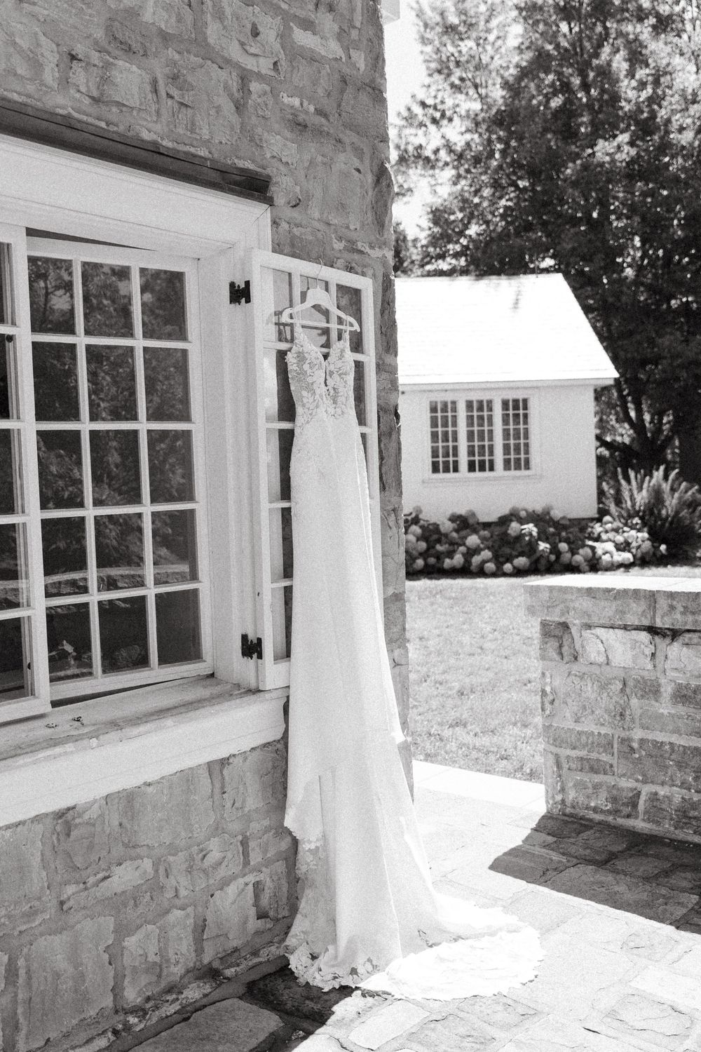 A delicate white wedding dress hangs elegantly against a stone wall exterior with French doors and garden views.
