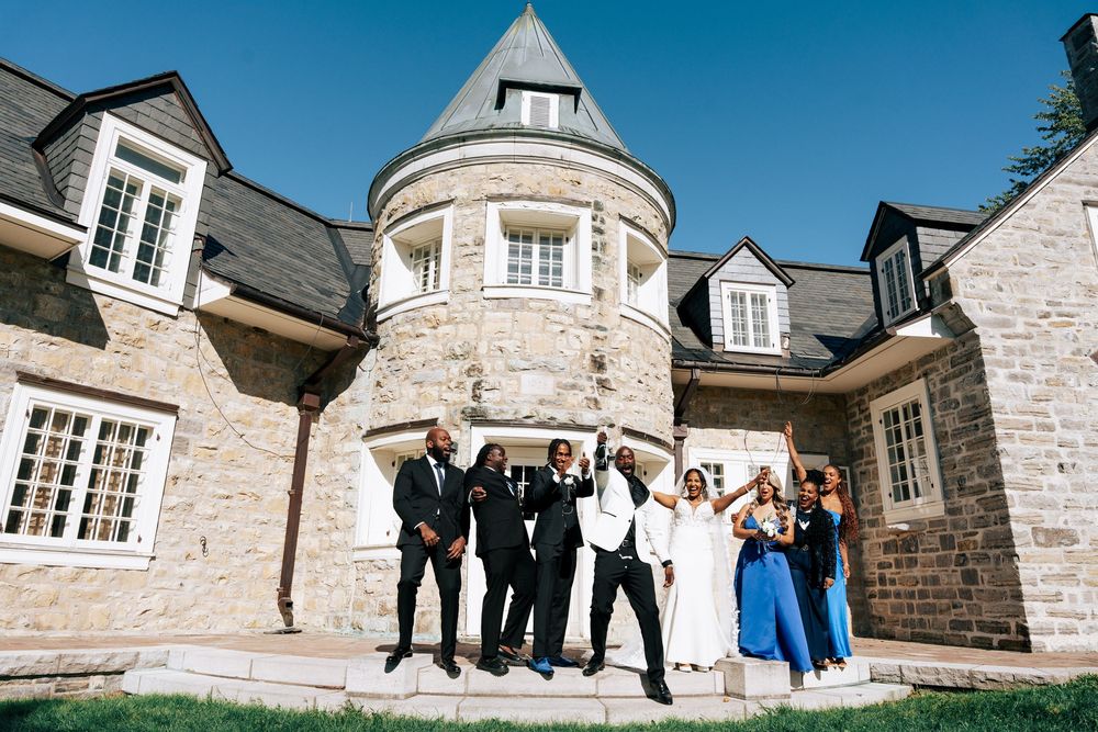 Wedding party members stand in front of a stone mansion with blue sky and architectural details visible.
