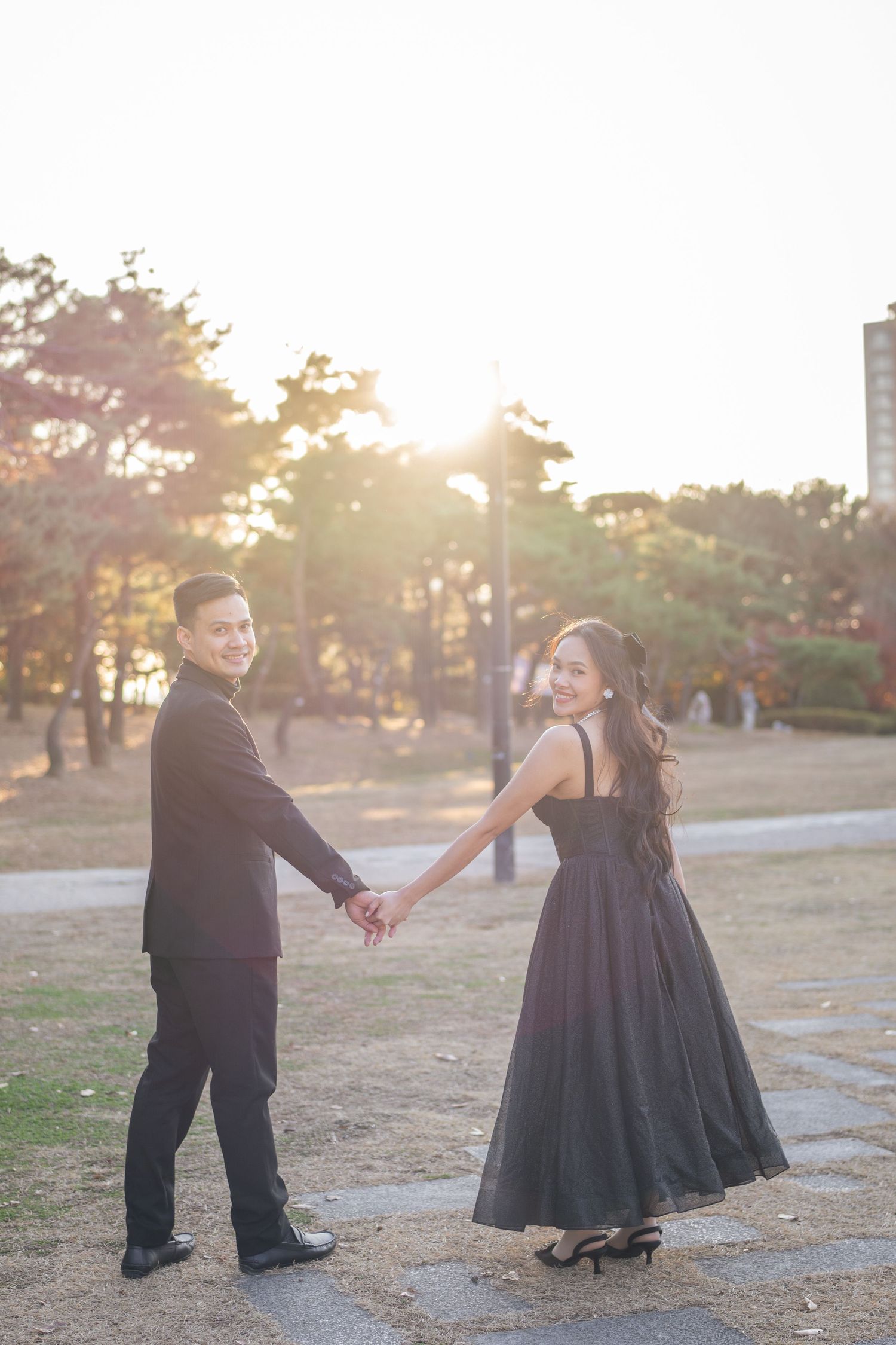 A couple in formal attire holds hands and poses in a park during golden hour sunset with trees in the background.