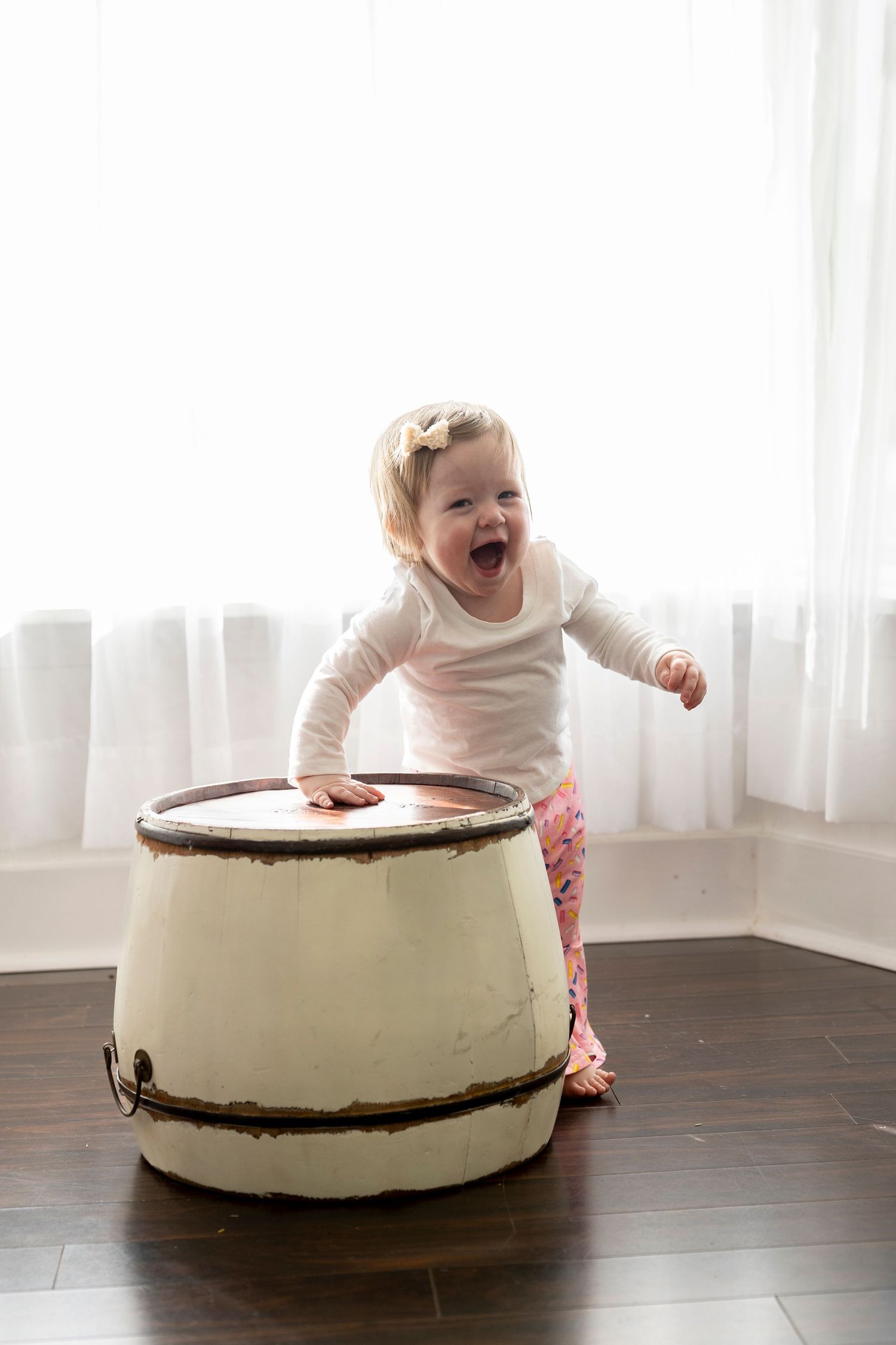A baby in white clothing plays and laughs while sitting next to a large round white cake on a dark wooden floor.