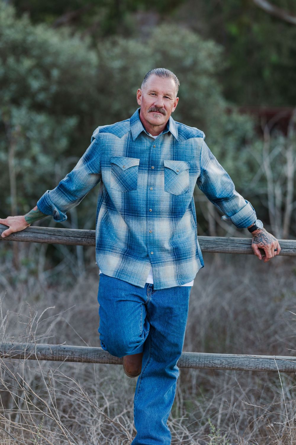 Person in plaid shirt and jeans poses casually against wooden fence in natural outdoor setting.