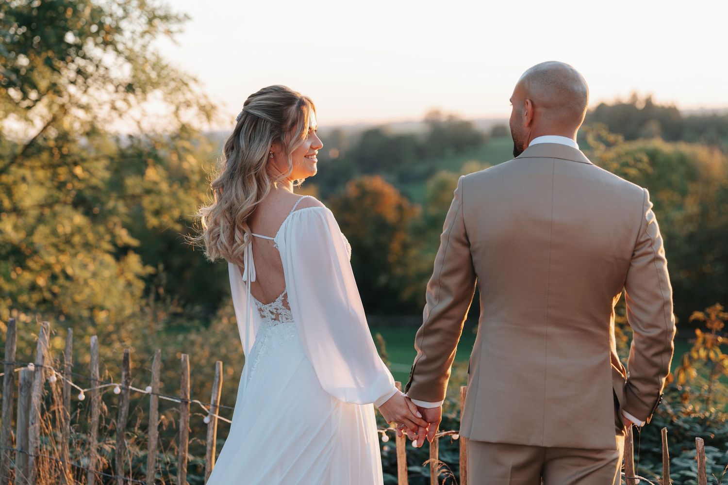 A couple in wedding attire stands together during golden hour in a scenic outdoor setting.
