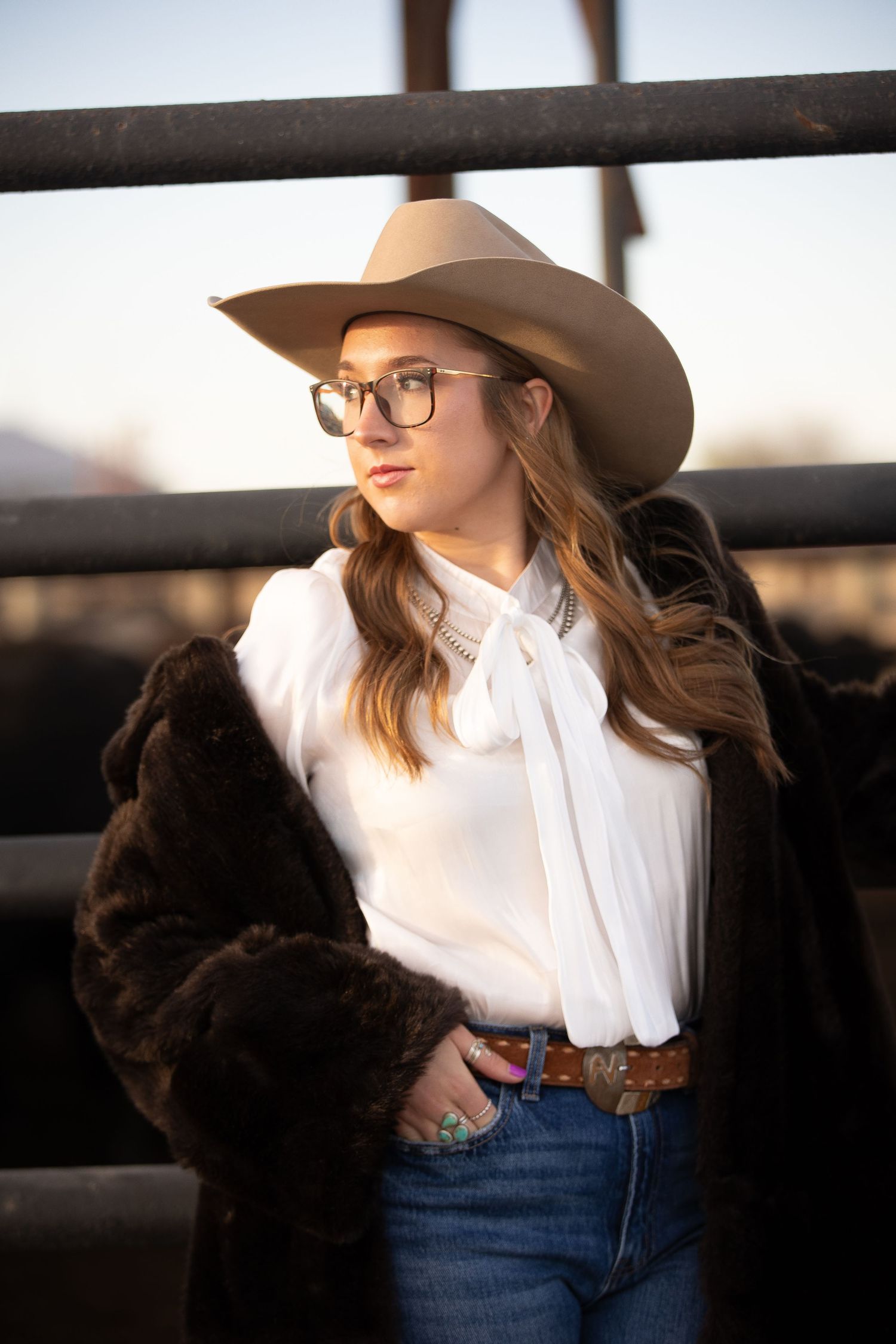 A person in a cowboy hat and white shirt leans against a rustic wooden fence at sunset.