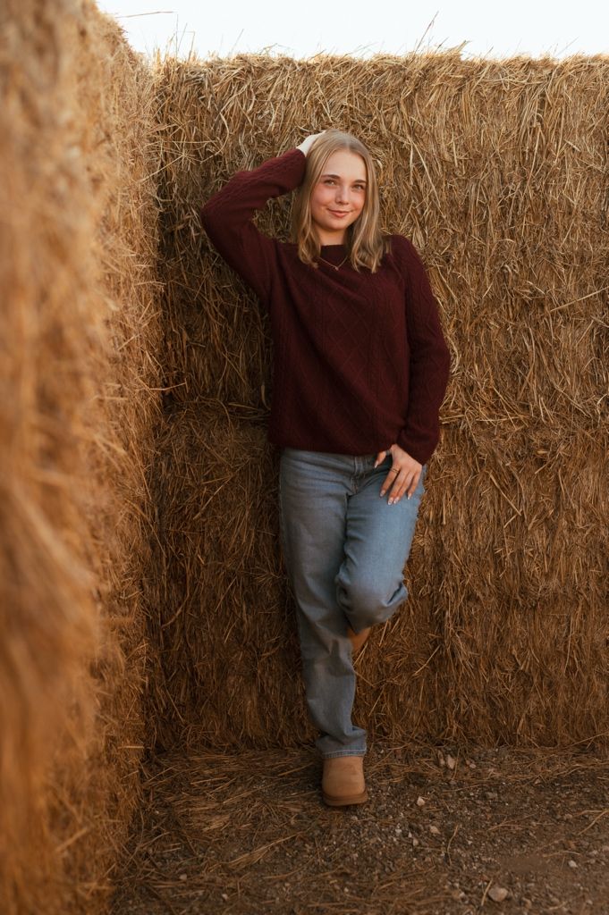 Person in burgundy sweater and jeans sits casually against straw bale backdrop in autumn setting.