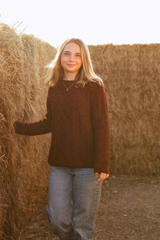 A person in a brown sweater and jeans poses against a golden hay bale backdrop during sunset.