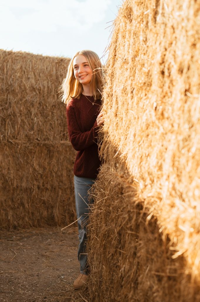 A person in a brown sweater stands against a golden hay bale wall illuminated by warm sunlight.