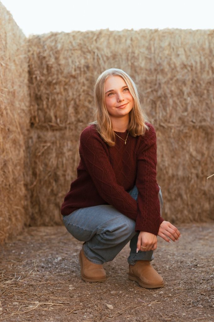 A casual autumn portrait against a rustic hay bale wall showing someone crouching in a burgundy sweater and jeans.