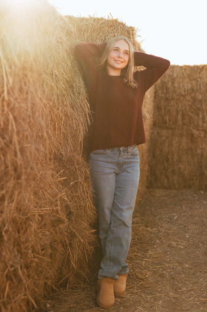 Person in jeans and brown boots poses against rustic hay bale wall in warm sunlit setting.
