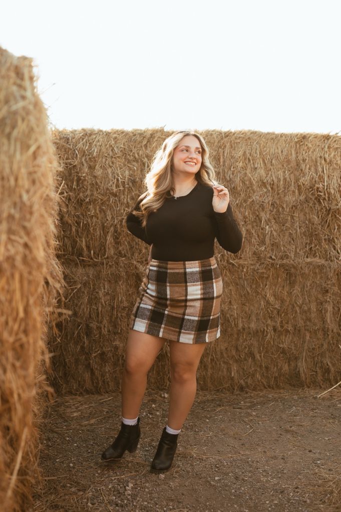 Person in black top and plaid skirt poses playfully by hay bale wall during golden hour.