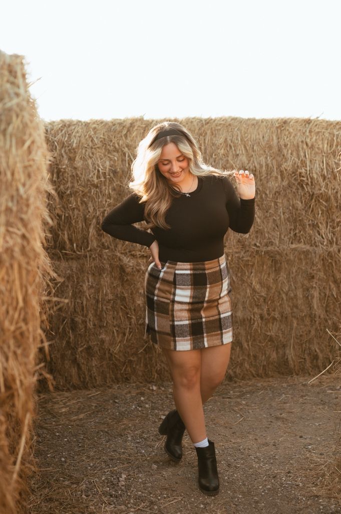 Person in black top and plaid skirt dances playfully against hay bale backdrop at sunset.